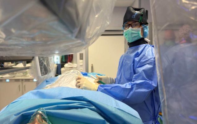Lekarz w sali zabiegowej wykonuje procedurę medyczną przy pacjencie leżącym na stole operacyjnym, przykrytym niebieskimi i zielonymi sterylnymi materiałami; pacjent ma maskę tlenową, a personel medyczny ubrany jest w niebieski fartuch, rękawice i maseczkę ochronną.