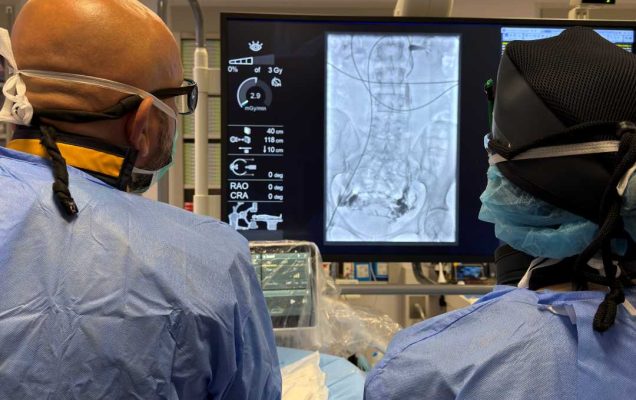 Dwóch lekarzy w sali zabiegowej obserwuje duży monitor z obrazem rentgenowskim przedstawiającym wnętrze ciała pacjenta; personel medyczny ubrany jest w niebieskie fartuchy, maski i czepki ochronne, a w tle widoczna jest aparatura medyczna.