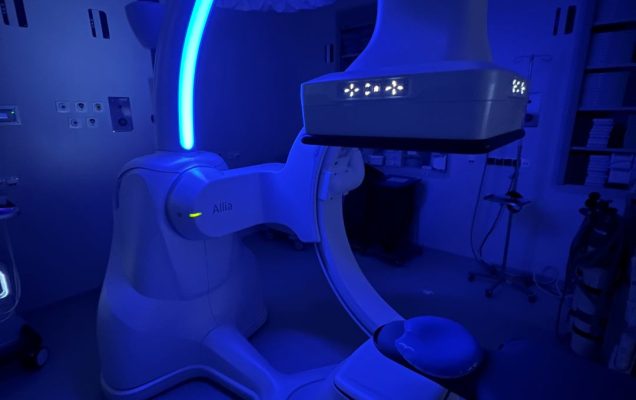 Nowoczesny aparat medyczny GE HealthCare typu Allia w sali zabiegowej, oświetlony niebieskim światłem; urządzenie ma charakterystyczne ramię w kształcie litery C i służy do obrazowania rentgenowskiego lub fluoroskopowego podczas procedur diagnostycznych.