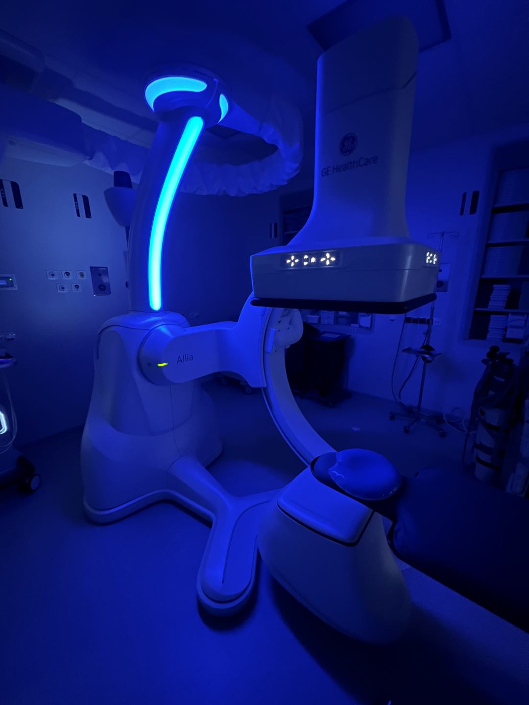 Nowoczesny aparat medyczny GE HealthCare typu Allia w sali zabiegowej, oświetlony niebieskim światłem; urządzenie ma charakterystyczne ramię w kształcie litery C i służy do obrazowania rentgenowskiego lub fluoroskopowego podczas procedur diagnostycznych.