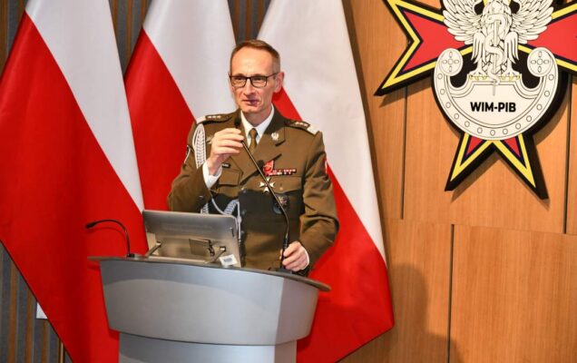 Grzegorz Gielerak przy mównicy. Za nim trzy flagi Polski i logotyp WIM.