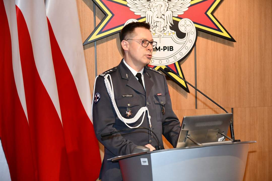 Jarosław Rytwiński przy mównicy. Za nim trzy flagi Polski i logotyp WIM.
