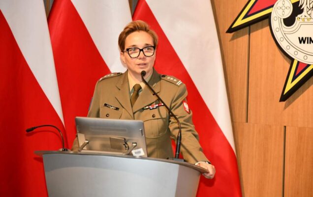 płk Anita Podlasin - Dyrektor Departamentu Wojskowej Służby Zdrowia przy mównicy. Za nią trzy flagi Polski i logotyp WIM.