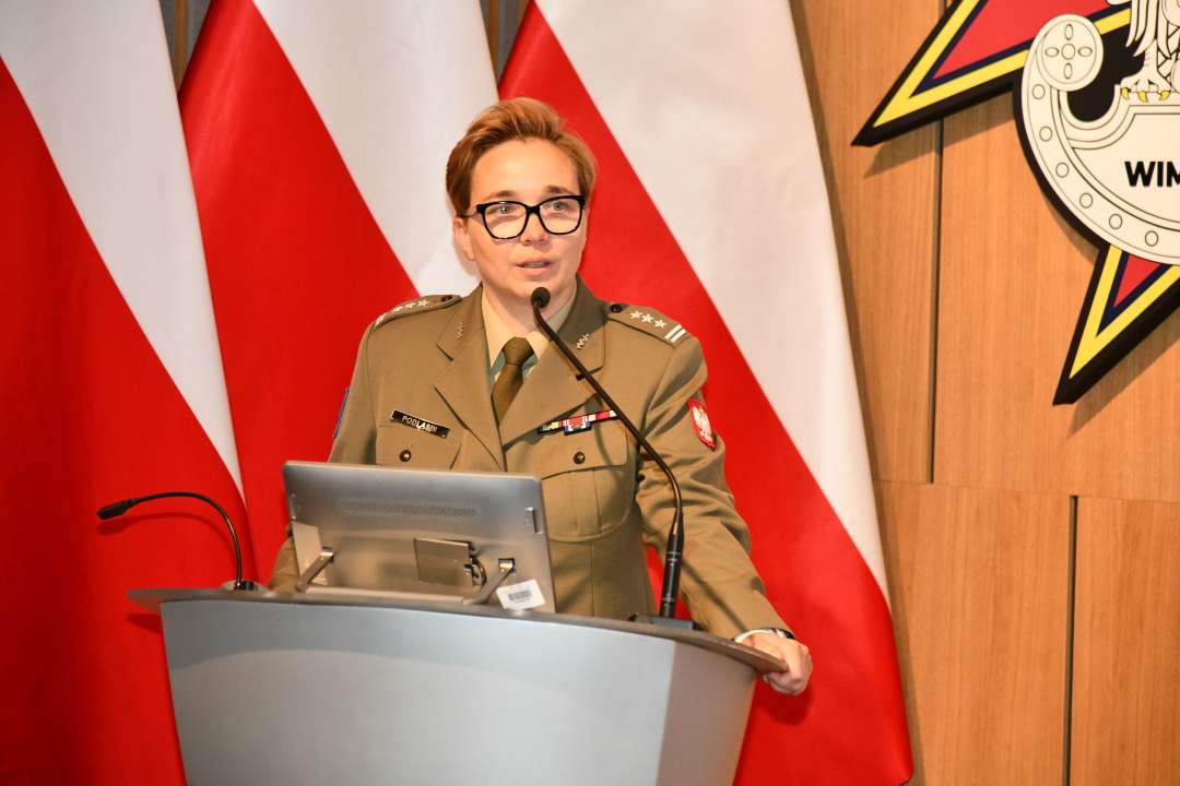 płk Anita Podlasin - Dyrektor Departamentu Wojskowej Służby Zdrowia przy mównicy. Za nią trzy flagi Polski i logotyp WIM.