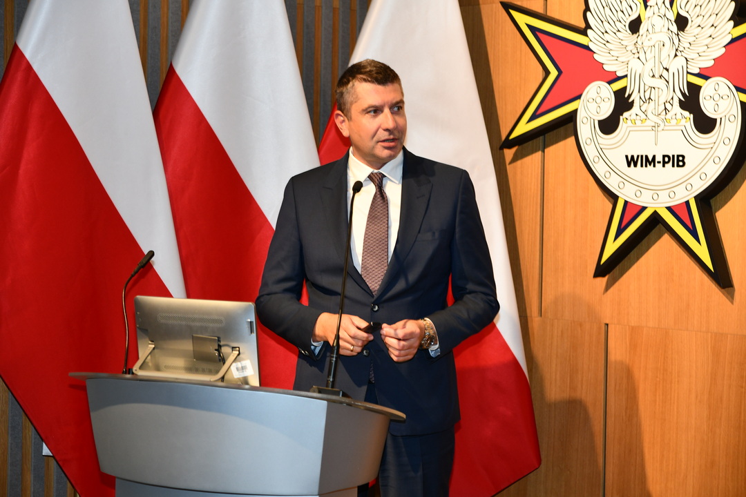 Adrian Szulczyński przy mównicy, za nim trzy flagi Polski oraz logo WIM