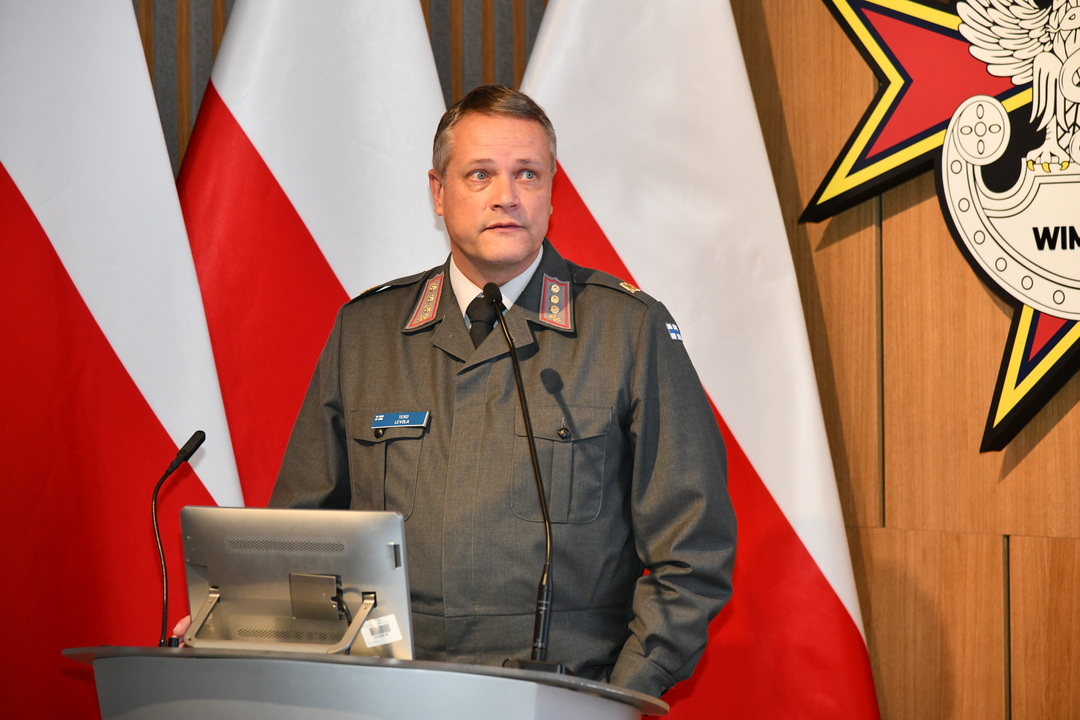 Gość Konferencji Tero Levola przy mównicy, za nim trzy flagi polski, oraz logo WIMu